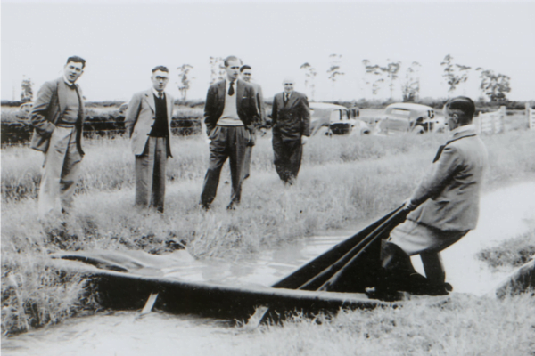The Duke of Edinburgh (third from left) observes border dyke irrigation at the Winchmore Research Station Irrigation Scheme. In a border dyke irrigation system, when water is diverted from the main water races into smaller ones, a temporary dam must be created at the outlet to each border, so the water spills through onto pasture. In this image the outlet behind the worker in the water has been closed with a board, the pasture on the right of it has been flattened by the previous flow of water. The worker is lifting a corner of the canvas dam to allow water to flow down to the next temporary dam.